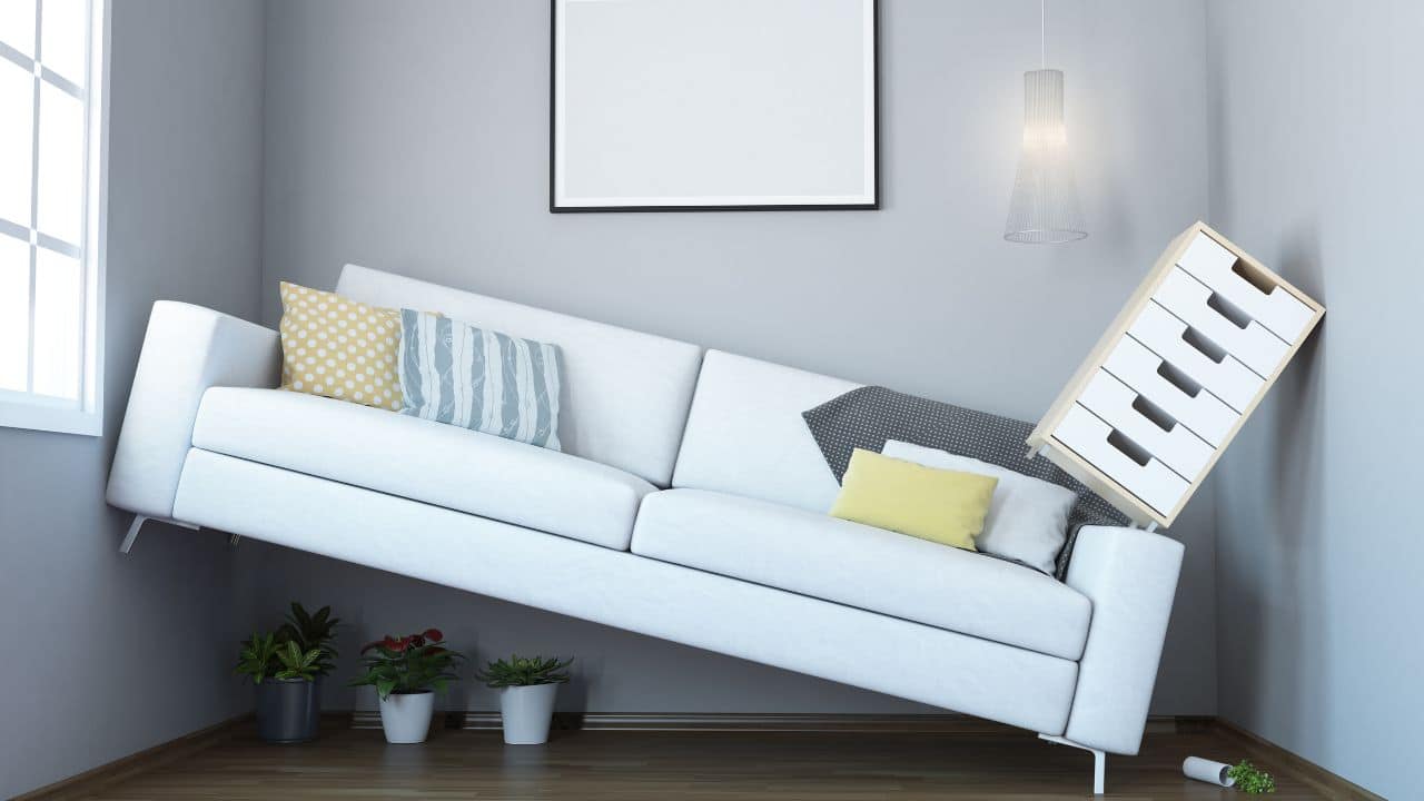 A white couch tilted at an angle with colorful throw pillows, a hanging light, and a leaning dresser, illustrating playful interior design hacks for visual impact.