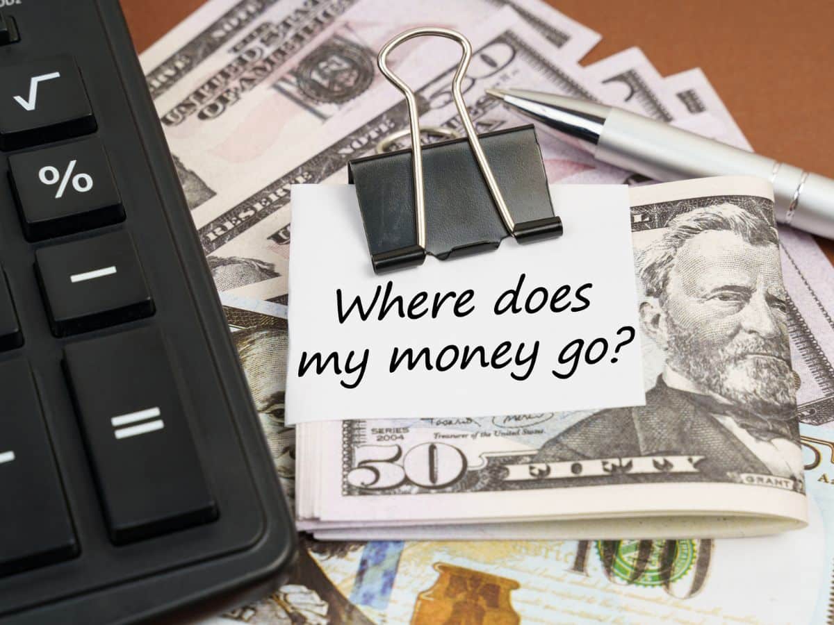 A stack of U.S. fifty-dollar bills sits under a note clipped with a binder that reads “Where does my money go?” next to a calculator and pen.